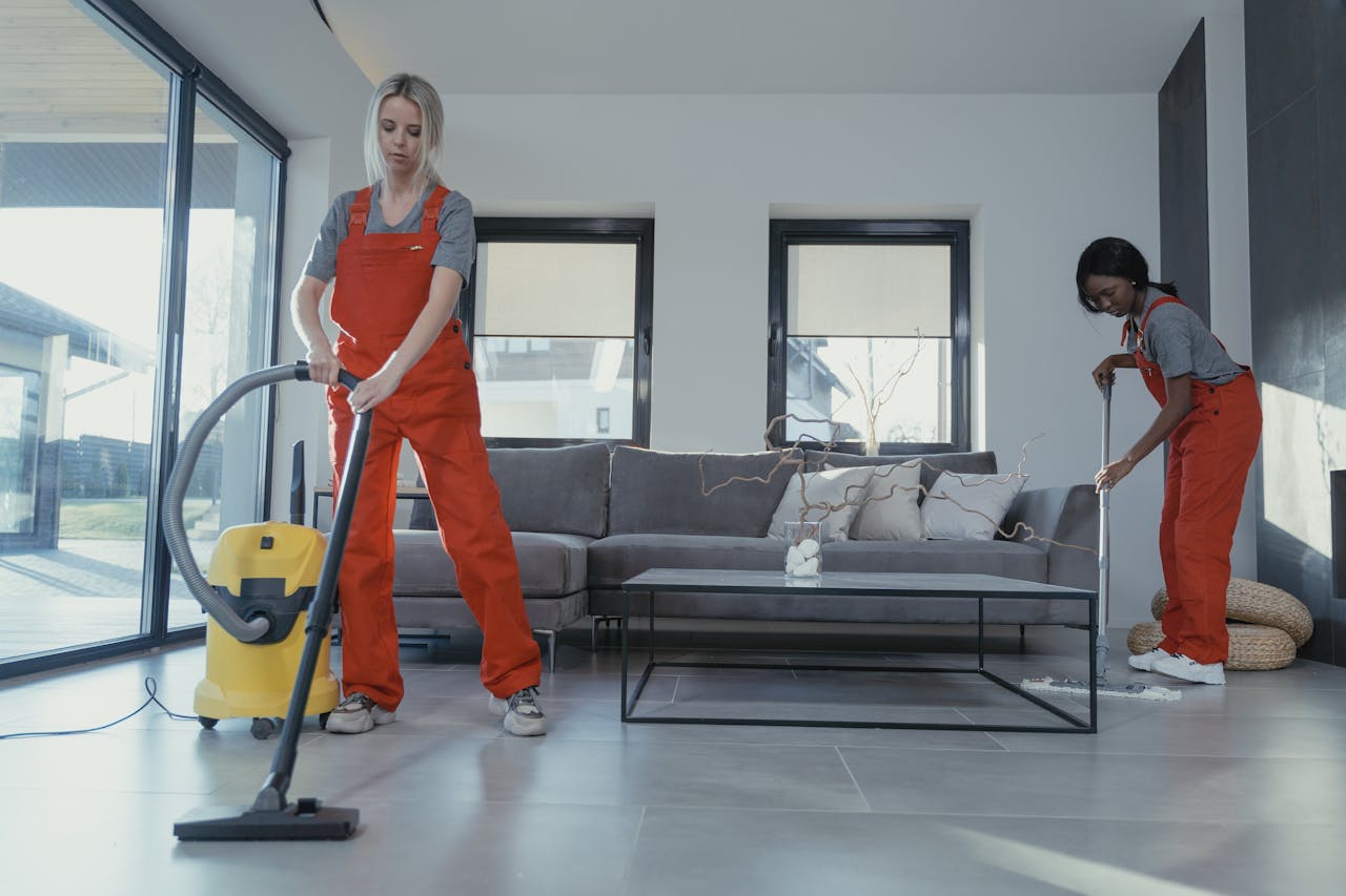 about-02 Two women in red overalls vacuuming and sweeping a modern living room.